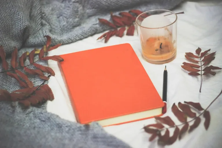 Plain orange book with a pen on a light blanket beside a candle and autumn leaves