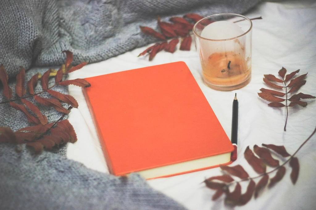 Plain orange book with a pen on a light blanket beside a candle and autumn leaves