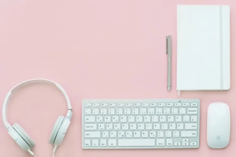 Pink desk flat lay with white headphones, a keyboard, a mouse, a notebook and a pen