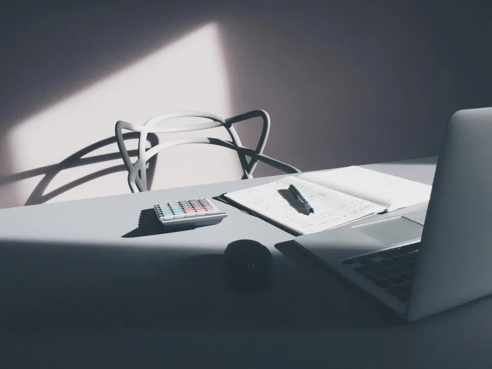 Minimal desk with an open notebook, pen, calculator and laptop in soft light