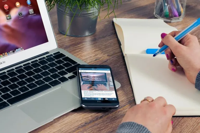 Laptop, smartphone and an open notebook on a wooden desk as someone takes notes