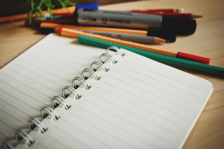 Open spiral notebook with blank lined pages next to colorful pens on a wooden desk