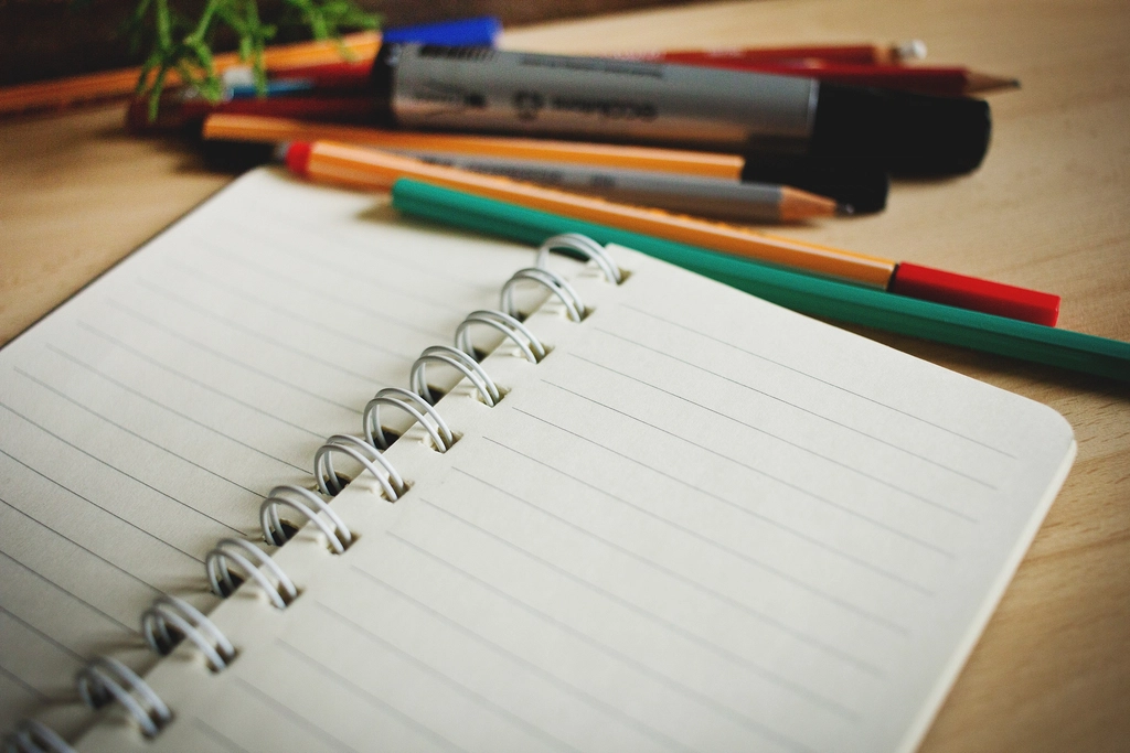 Open spiral notebook with blank lined pages next to colorful pens on a wooden desk