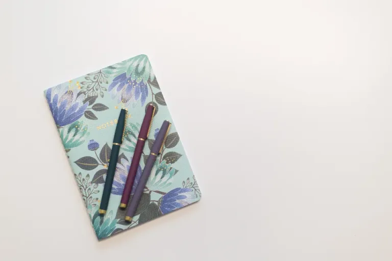 A floral-covered notebook with three pens on a white table