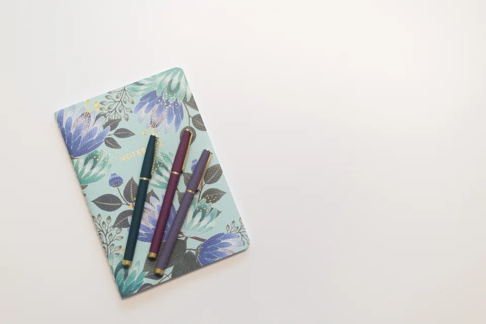 A floral-covered notebook with three pens on a white table