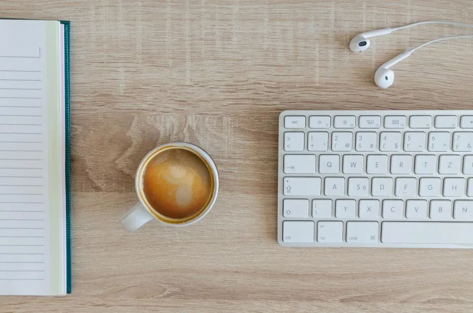 Wooden desk with a keyboard, a cup of coffee, earphones and an open notebook seen from above