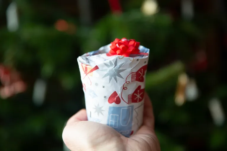 Hand holding a small wrapped holiday gift with a red bow in front of a decorated tree